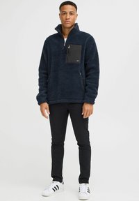 Navy fleece pullover with a black zippered chest pocket, high collar, and textured surface, paired with black pants and white sneakers.