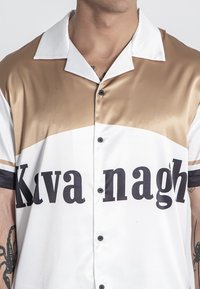 Camisa de botón de manga corta con un diseño brillante en oro y blanco, que presenta letras negras en negrita y acentos a rayas en contraste en las mangas.