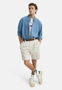 Camisa de mezclilla azul claro, camiseta gráfica blanca debajo, pantalones cortos beige, cinturón marrón, zapatillas blancas con detalles verdes y calcetines a rayas.