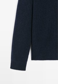 Manica di un maglione a coste in navy con una consistenza morbida. I polsini presentano un orlo dritto sottile e uno schema coerente in tutto il capo.