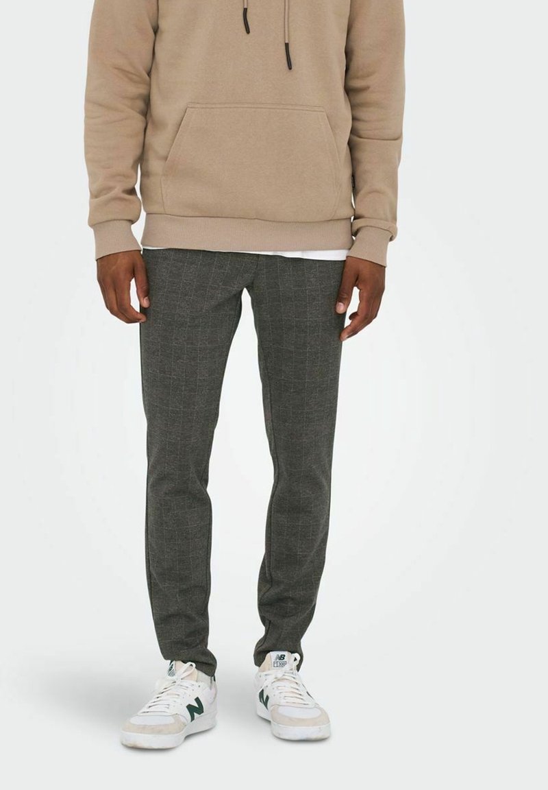 Lichtbeige hoodie met een voorzak, gecombineerd met donkergrijs geruite tapered pants en witte sneakers met groene accenten.