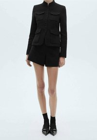 Femme portant une veste noire à boutons avec quatre poches à rabat, des shorts noirs, des chaussettes noires et des chaussures à bout pointu noires sur un fond clair.