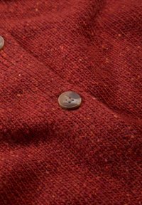 Detail z blízka na pletenou látku v rezavé barvě s lesklým hnědým knoflíkem přišitým na ní, ukazující podrobné textury a stehy.