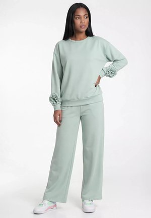 Kobieta w jasnozielonych szerokich spodniach i bluzie z kwiatowymi detalami na rękawach, nosząca białe trampki, stoi z ręką na biodrze.