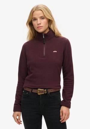 Pull en polaire bordeaux avec un col zippé quart, doté de manches longues et d'un petit logo sur la poitrine. Porté avec un jean noir et une ceinture marron.