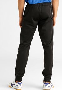 Pantalons de sport noirs avec taille élastique et ourlets resserrés. Texture lisse avec une coupe ample. Portés avec des chaussures de sport blanches et bleues.