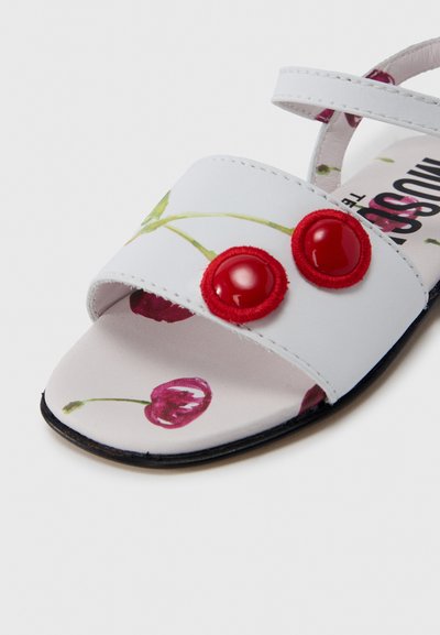 Λευκό σανδάλι με λουλουδάτο σχέδιο και κόκκινα υφασμάτινα κουμπιά. Ανοιχτό σχέδιο στα δάχτυλα, επίπεδη σόλα και ρυθμιζόμενα λουράκια για ασφαλή εφαρμογή.