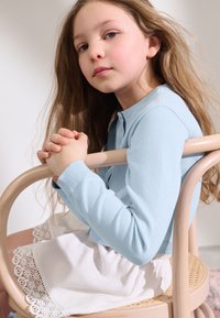 Light blue cardigan with a round neck, paired with a white skirt featuring lace trim. Sitting on a wooden chair, with textured hair.