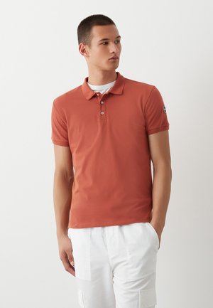Polo arancione ruggine con colletto classico, patta con due bottoni e maniche corte. Presenta una toppa con logo sulla manica sinistra.