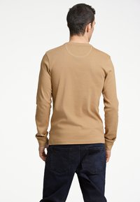 Långärmad skjorta i en beige färg, tillverkad av ett mjukt bomullstyg. Har en rundad halsringning och åtsittande manschetter, kombinerad med mörka jeans.