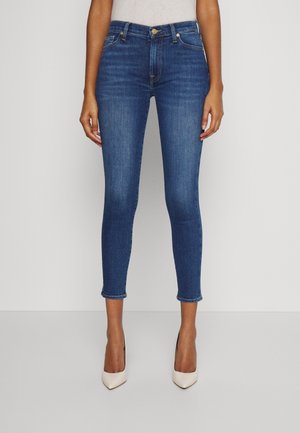 Frau trägt schlanke, knöchellange blaue Jeans mit leichter Verblassung an den Oberschenkeln und weißen High-Heels, steht vor einem schlichten Hintergrund.