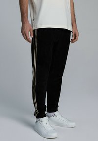 Czarne spodenki dresowe z kreszami w kolorze kremowym wzdłuż boków, zwężany krój, wykończone ściągaczem na kostkach, połączone z białymi sneakersami.