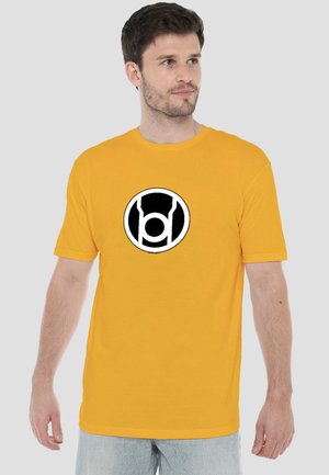 Junger Mann trägt ein gelbes T-Shirt mit einem schwarz-weißen kreisförmigen grafischen Symbol auf der Brust und steht vor einem schlichten hellen Hintergrund.