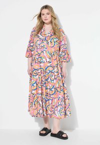 Farbenfrohes gemustertes Kleid mit einer Mischung aus pinken, blauen und gelben Farbtönen, das mit gestuften Rüschen und drei Viertelärmeln versehen ist. Kombiniert mit schwarzen Sandalen.