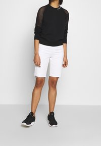 Czarna bluza z siateczkowymi rękawami, zestawiona z białymi bermudami. Model nosi czarne buty sportowe z białymi podeszwami.