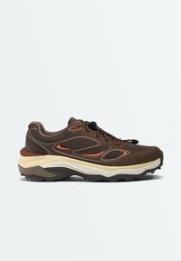 Baskets de sport marron avec des accents orange, semelle beige et noire épaisse, et fermeture élastique à lacets sur un fond clair uni.