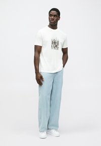 Hvit bomull T-skjorte med grafisk print i midten. Modellen har på seg lyseblå vide jeans og hvite sneakers.
