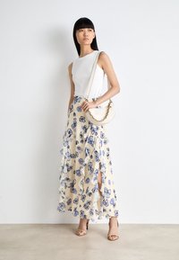 Top bianco senza maniche abbinato a una gonna maxi gialla a fiori con motivi blu e volant, completata da una borsa beige e tacchi.