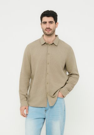 Άνδρας με σκούρα μαλλιά, ντυμένος με μπεζ πουκάμισο με μακριά μανίκια και ανοιχτό μπλε τζιν, στέκεται με το ένα χέρι στην τσέπη.