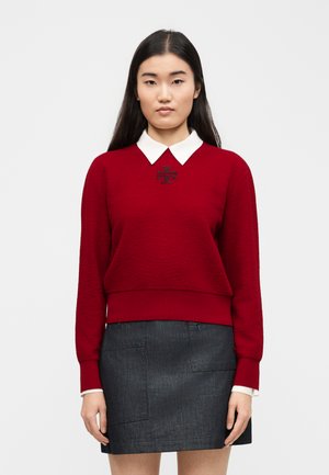 Γυναίκα που φορά κόκκινο πουλόβερ με λευκό γιακά και μαύρο λογότυπο, συνδυασμένο με μίνι φούστα από σκούρο τζιν, στέκεται μπροστά σε απλό φόντο.