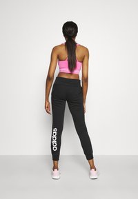 Schwarze Jogginghose mit weißem "adidas"-Logo am linken Bein, kombiniert mit einem pinkfarbenen Sport-BH. Das Model steht mit dem Rücken zur Kamera und hat lange Haare.