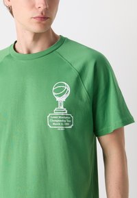 Žalias medvilninis marškinėlis su apvaliu iškirpimu. Pavaizduotas baltas trofėjus ir tekstas: "Žemutinio Manhatano čempionato turas 1990 m. kovo 11 d."