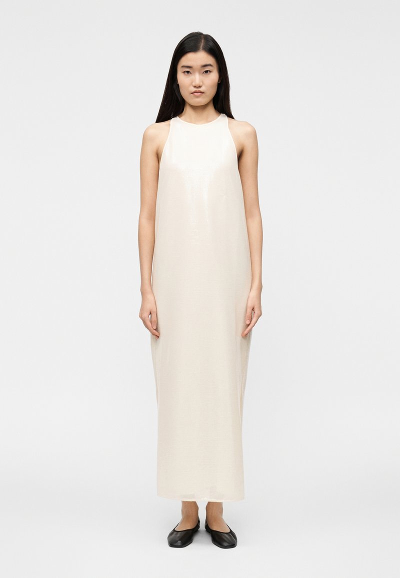 Μάξι φόρεμα αμάνικο σε κρεμ απόχρωση με ροή στη σ ιλουέτα. Λείο υλικό, μινιμαλιστικός σχεδιασμός και διακριτική λάμψη. Συνδυάζεται με μαύρες μπαλαρίνες.