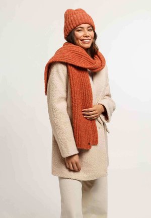 Un bonnet et une écharpe en tricot côtelé orange associés à un manteau crème texturé, avec des détails de boutons et des manches décontractées, offrant chaleur et style.