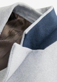 Abrigo de lana gris con un cuello azul marino. El forro interior es marrón con un patrón sutil. Tejido texturizado y un diseño de cuello limpio y ajustado.