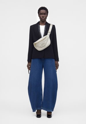 Γυναίκα που φοράει μαύρο σακάκι, λευκό τοπ, φαρδιά μπλε τζιν, μαύρα παπούτσια και μπεζ τσάντα crossbody με μεγάλο λογότυπο στο μπροστινό μέρος.