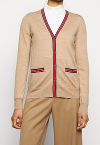 Beige Strickjacke mit V-Ausschnitt, roten und marineblauen Akzenten, zwei Vorder pockets und dekorativen Knöpfen. Glatte Textur und taillierte Passform.