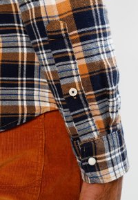 Fleedtröjan har ett rutigt mönster i marinblått, orange och vitt, med en knappstängning på ärmslutet. Tyget verkar mjukt och strukturerat.