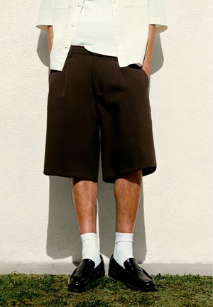 Person steht an einer weißen Wand und trägt schwarze lange Shorts, weiße Socken, schwarze Loafers, ein weißes kurzärmeliges Hemd und ein weißes Unterhemd.