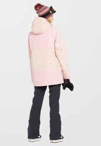 Helle rosa, farblich abgesetzte Außenjacke mit Kapuze, kombiniert mit schwarzen Hosen und Handschuhen, getragen mit einer weinroten Mütze mit Pompon.