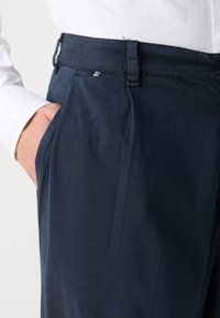 Ναυτικό μπλε παντελόνι με λεία υφή, που διαθέτει ζώνη με θηλιές και μια διακριτική μεταλλική λεπτομέρεια στο μπροστινό μέρος.