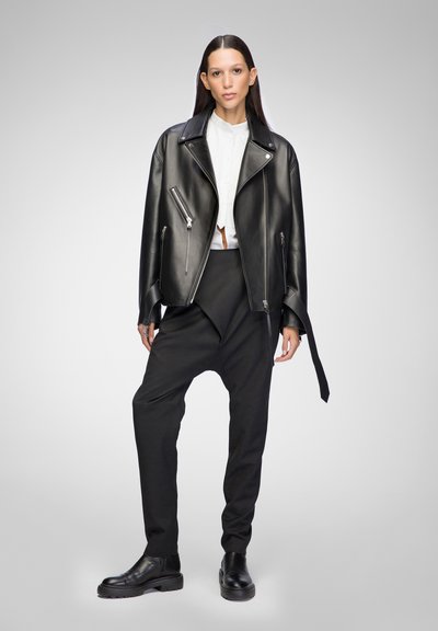 Schwarze Leder-Bikerjacke mit silbernen Reißverschlüssen, kombiniert mit lockeren schwarzen Hosen und schwarzen Stiefeletten. Weißes Hemd unter der Jacke.
