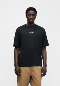 Μαύρο βαμβακερό μπλουζάκι με κοντά μανίκια και στρογγυλή λαιμόκοψη. Στο στήθος φαίνεται ένα μπλε λογότυπο. Συνδυάζεται με ανοιχτό καφέ παντελόνια.