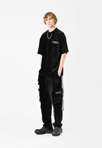 Camisa de corto con cuello de pana negra con bolsillo en el pecho y detalle de cadena, combinada con pantalones cargo de pana negra que cuentan con múltiples bolsillos y acentos.