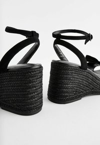 Sorte kile-sandaler med en tekstureret, flettet jute-platform og sorte ruskindssnore. Har ankel-lukning og glat inderside.