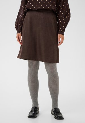 Vrouw draagt een bruine rok, een bruine blouse met stippen, grijze geribde panty's en zwarte glanzende loafers, staand op een witte achtergrond.