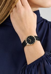 Črna ročna ura z okroglo številčnico, zlatimi detajli in črnim usnjenim trakom. Ura se ponaša z minimalističnim oblikovanjem z tankimi oznakami ur.