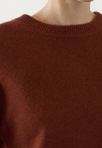 Pull en tricot couleur rouille avec un col rond à côtes, présentant une texture douce et un léger éclat. La vue en gros plan montre le détail du tissu.