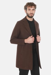 Cappotto in misto lana marrone con rever a punta, design monopetto e tasche laterali, indossato sopra un maglione nero texture.