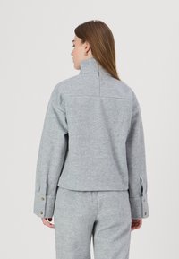 Grå fleecejakke med høy krage, drop shoulders og knappede mansjett-detaljer. Stoffet har en myk, teksturert utseende.