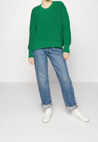 Pull en maille côtelée verte avec un col rond, porté sur un jean bleu large, associé à des sneakers blanches.