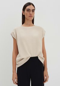 Top oversize lavorato a maglia beige con maniche corte, scollo rotondo e dettaglio con nodo laterale, abbinato a pantaloni neri. Texture liscia.