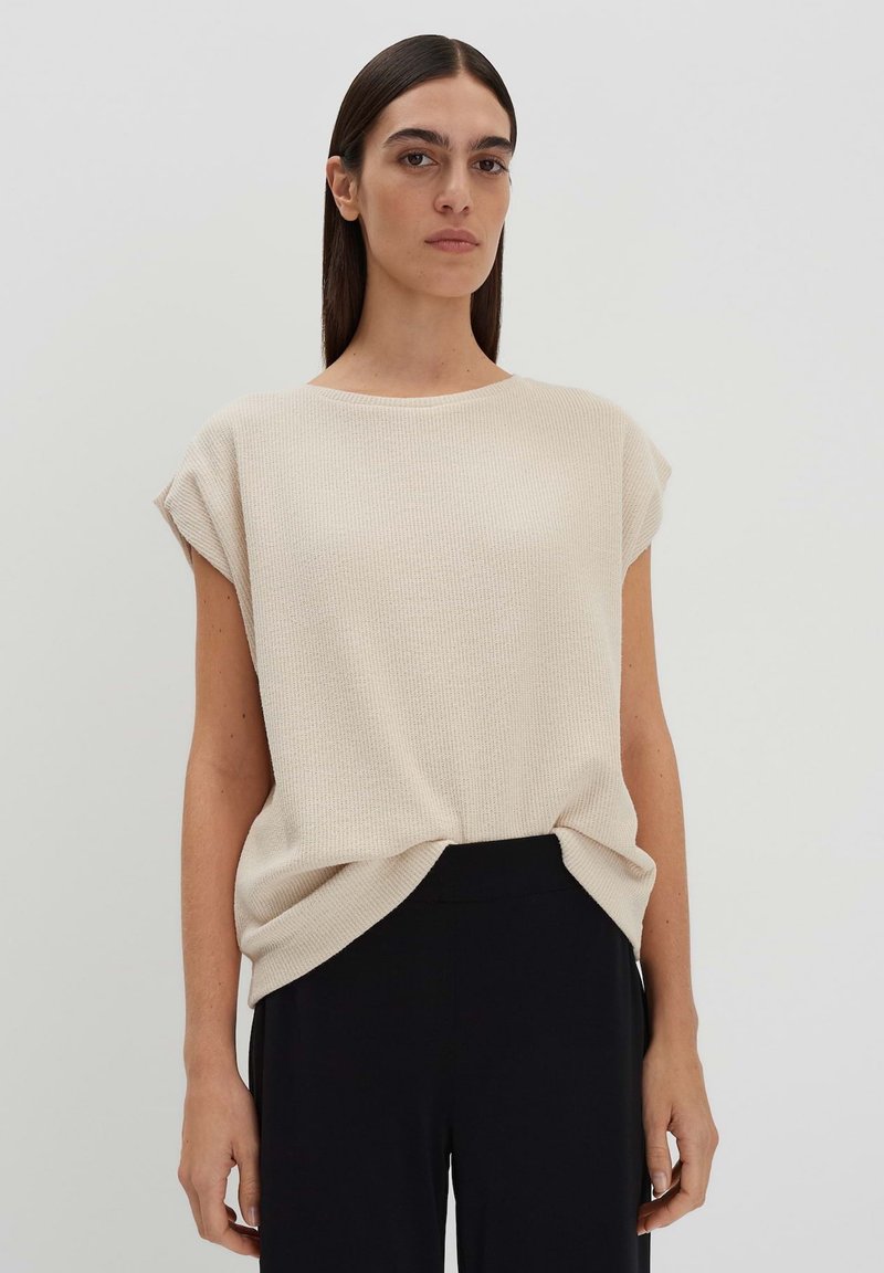 Top oversize lavorato a maglia beige con maniche corte, scollo rotondo e dettaglio con nodo laterale, abbinato a pantaloni neri. Texture liscia.