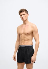 Modèle masculin aux cheveux courts et bouclés portant un boxer Lacoste noir sur un fond gris clair uni.