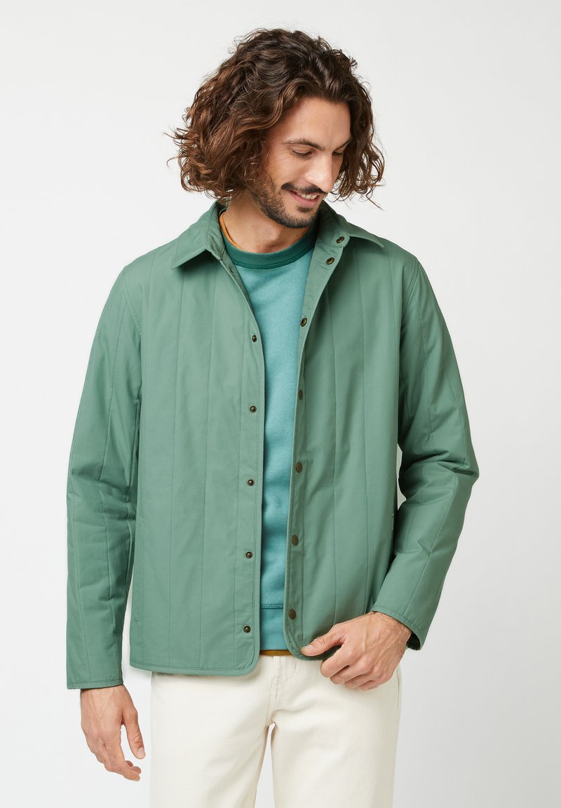 Groene, gewatteerde jas met een kraag, drukknoopsluiting en lange mouwen. Gedragen over een blauwe trui, met een gladde, lichte textuur.
