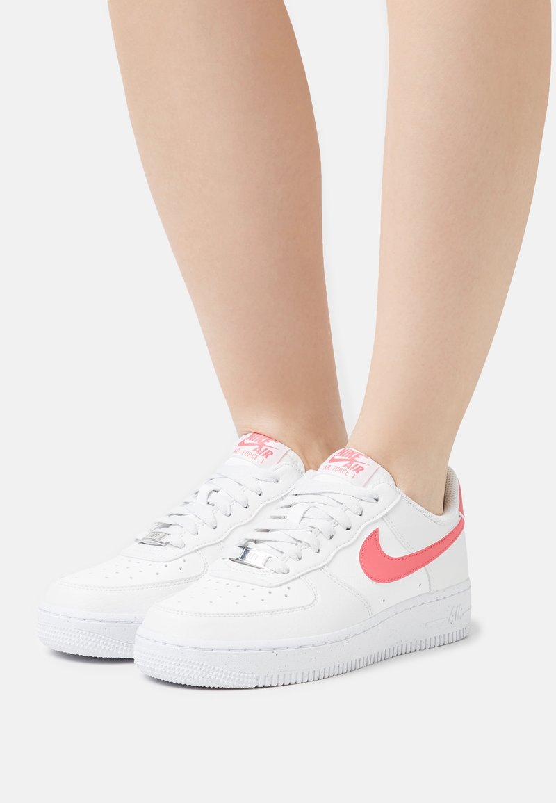 Baskets en cuir blanc avec un swoosh et un marquage Nike roses. La semelle est texturée avec des rainures ; bout rond et design à lacets.
